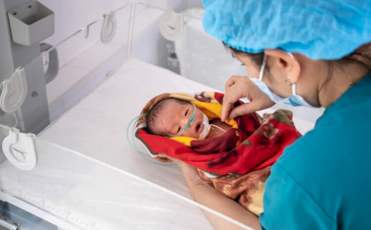 A pre-term infant of Asian descent is placed into a medical crib by a medical professional wearing scrubs and a mask. The infant is wrapped in a red and yellow cloth and has an oxygen tube attached to their nose.